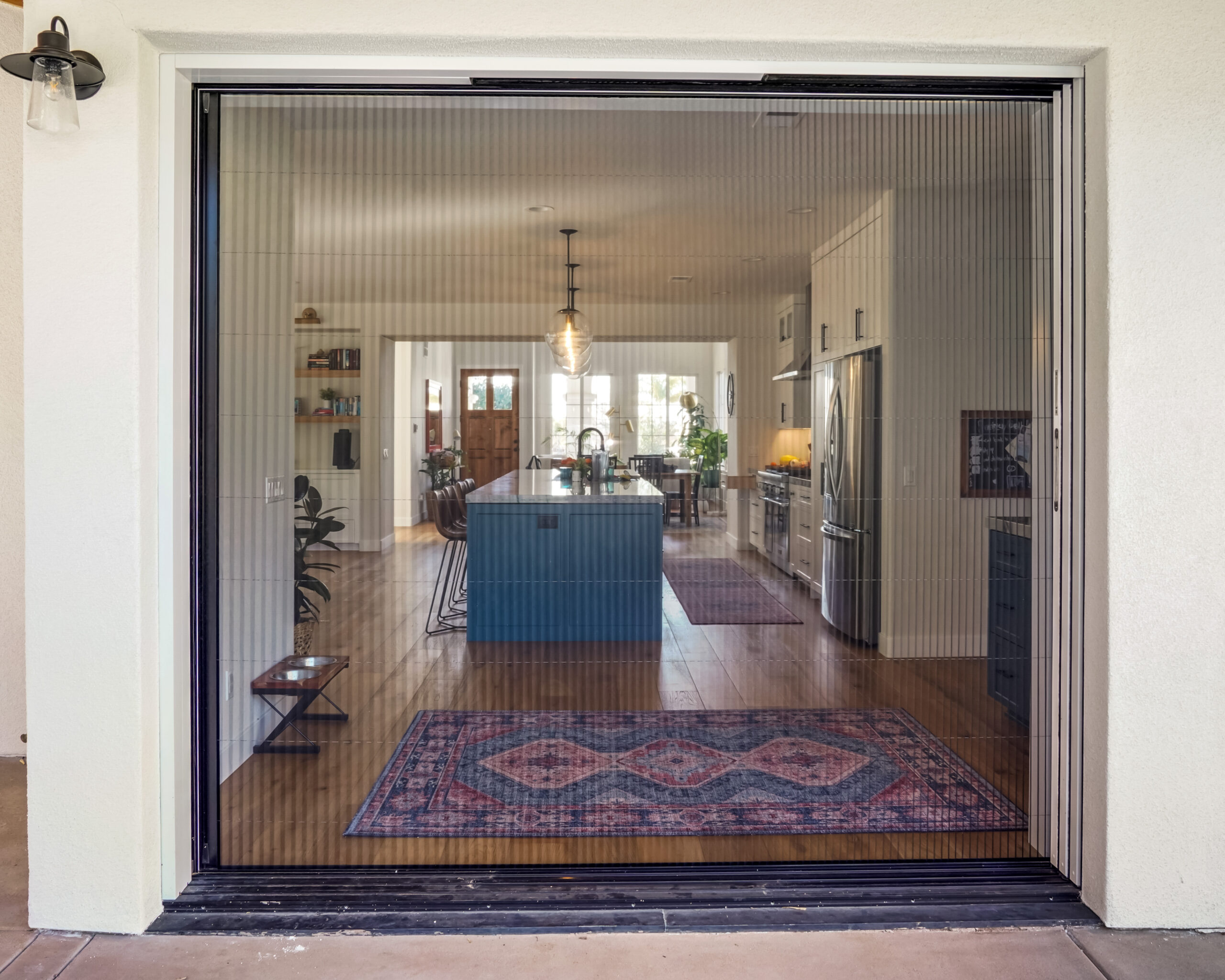Untitled-465 A modern kitchen with a large island is visible through a transparent closed sliding door with vertical blinds. The view also shows wooden flooring and decorative rugs.
