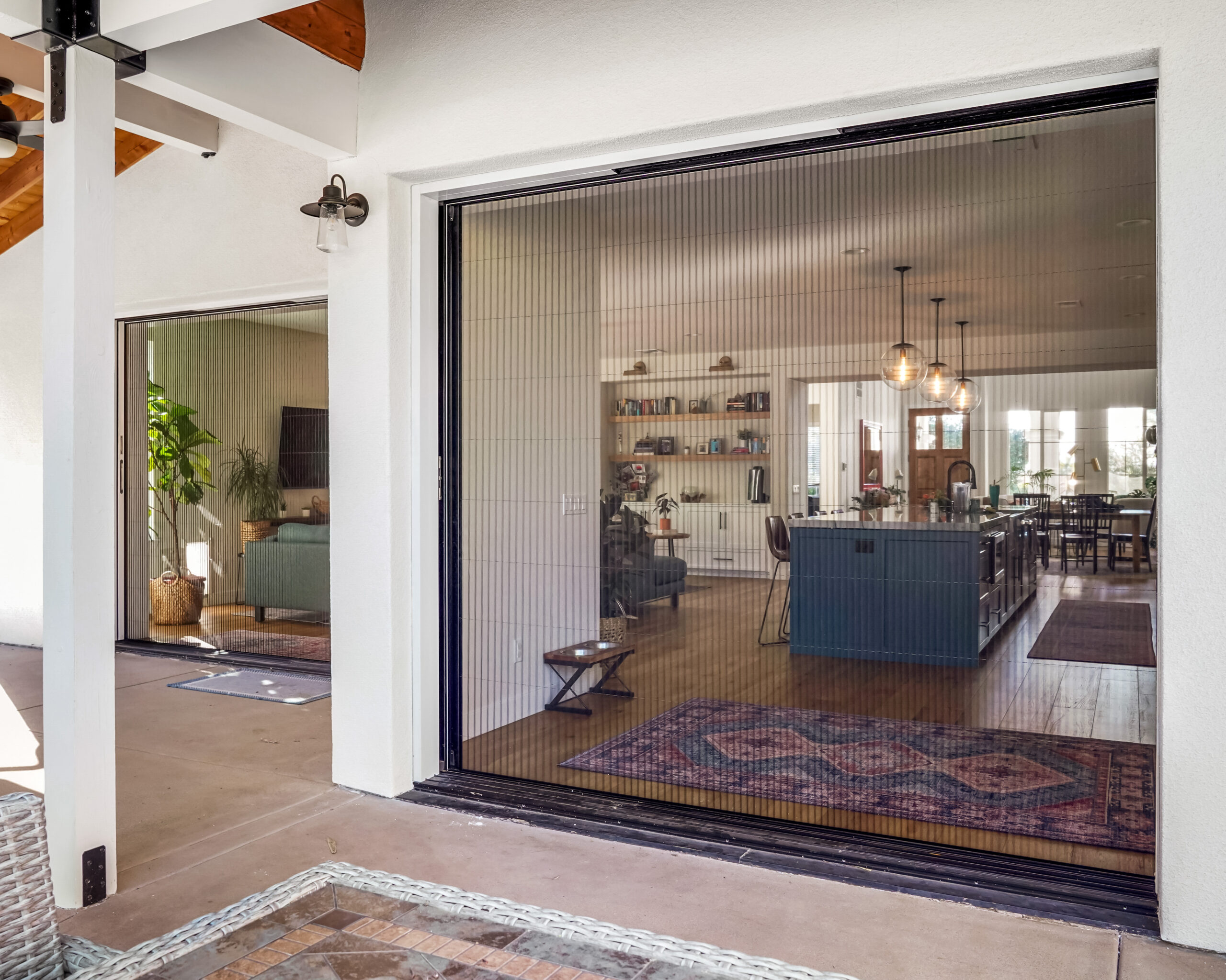 Untitled-466 A modern living room and kitchen are visible through large, open sliding glass doors from a covered patio area. The interior has minimalist decor with a bookshelf, plants, and pendant lights.