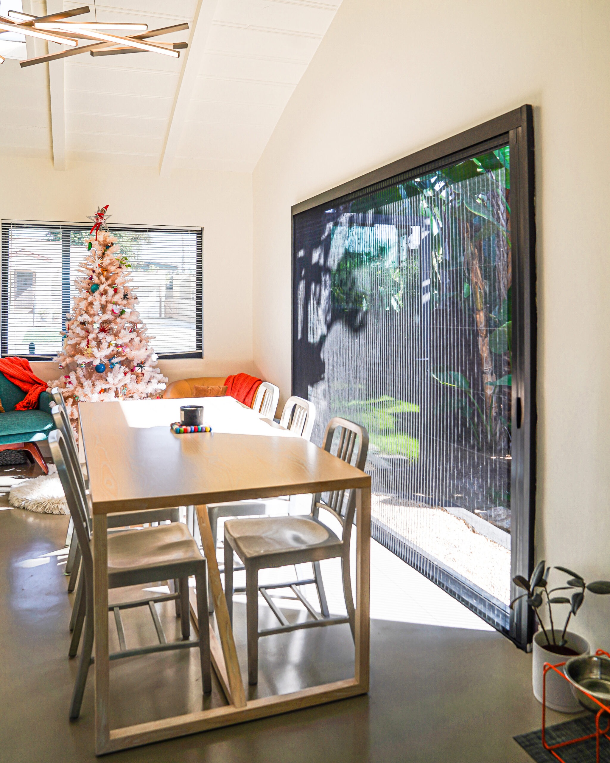 Untitled-468 A dining room with a wooden table and metal chairs, a white Christmas tree, a large window with vertical blinds, and potted plants.