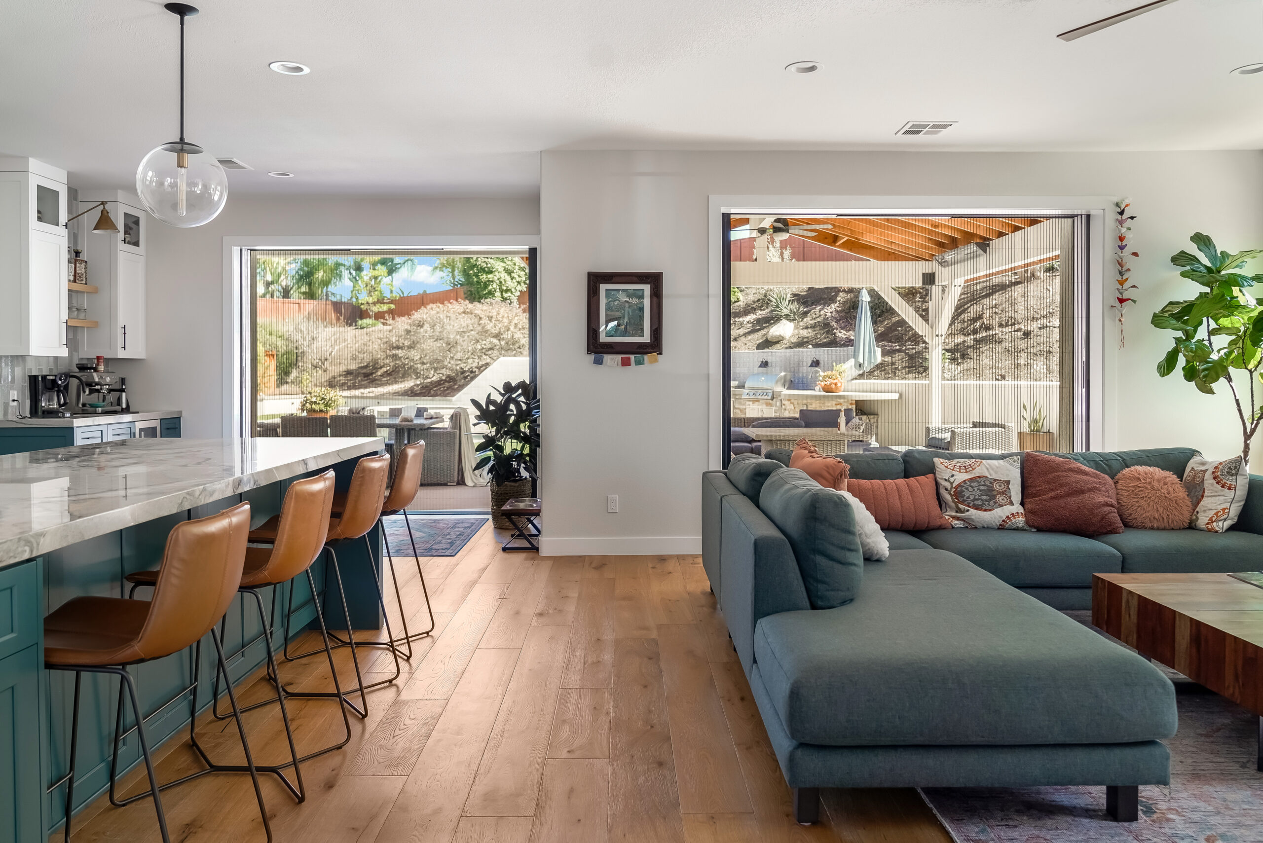 Untitled-470 A modern living room featuring an L-shaped sectional sofa, adjacent to an open kitchen with a marble-topped island and brown barstools. Large sliding doors provide access to a patio area outside.