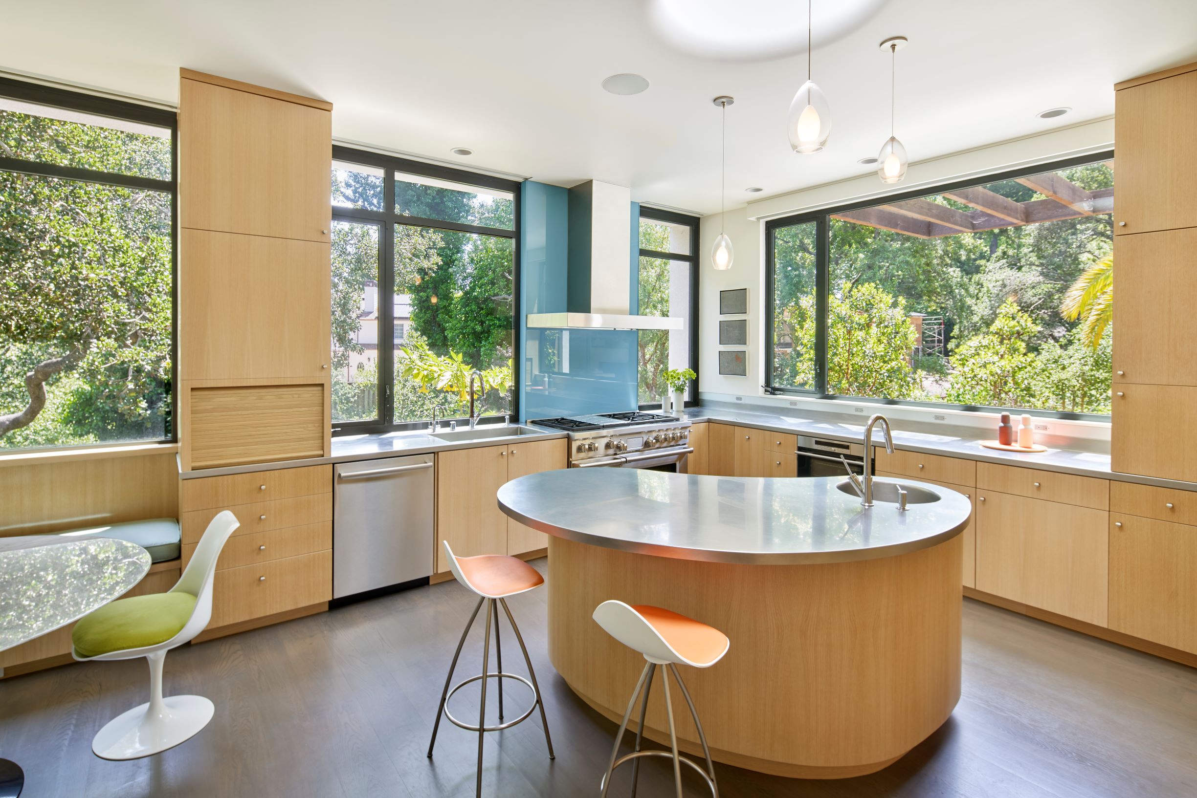 Modern kitchen with light wood cabinets, a curved island with stools, stainless steel appliances, large windows, and a built-in breakfast nook overlooking a green outdoor view.